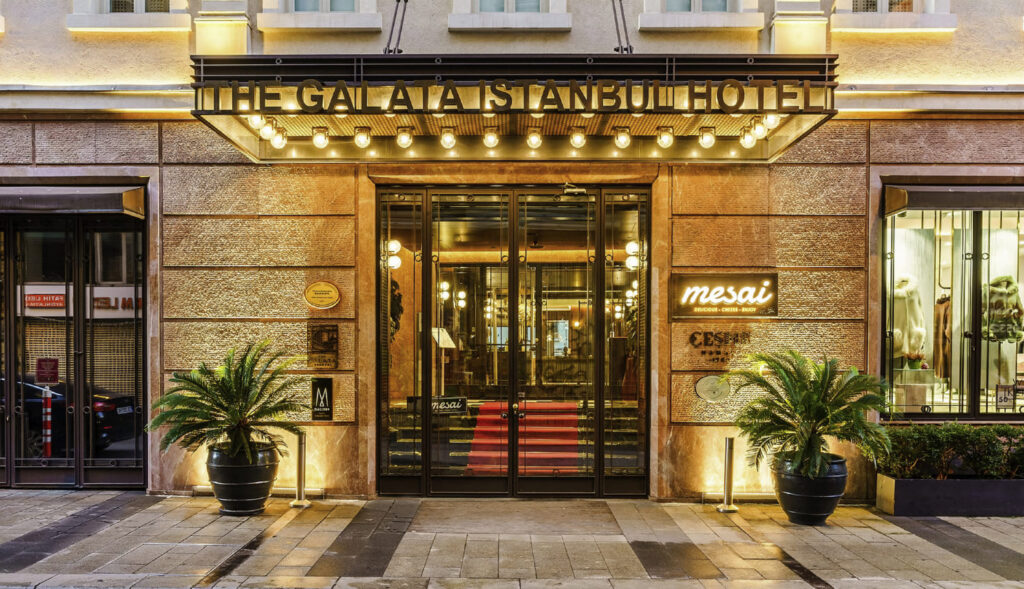 The Galata Istanbul Hotel MGallery illuminated street entrance at night on Bankalar Caddesi, with brass lettered marquee sign, vintage globe lights, glass doors with red carpet interior, flanking palm planters, and Mesai restaurant neon sign to the right
