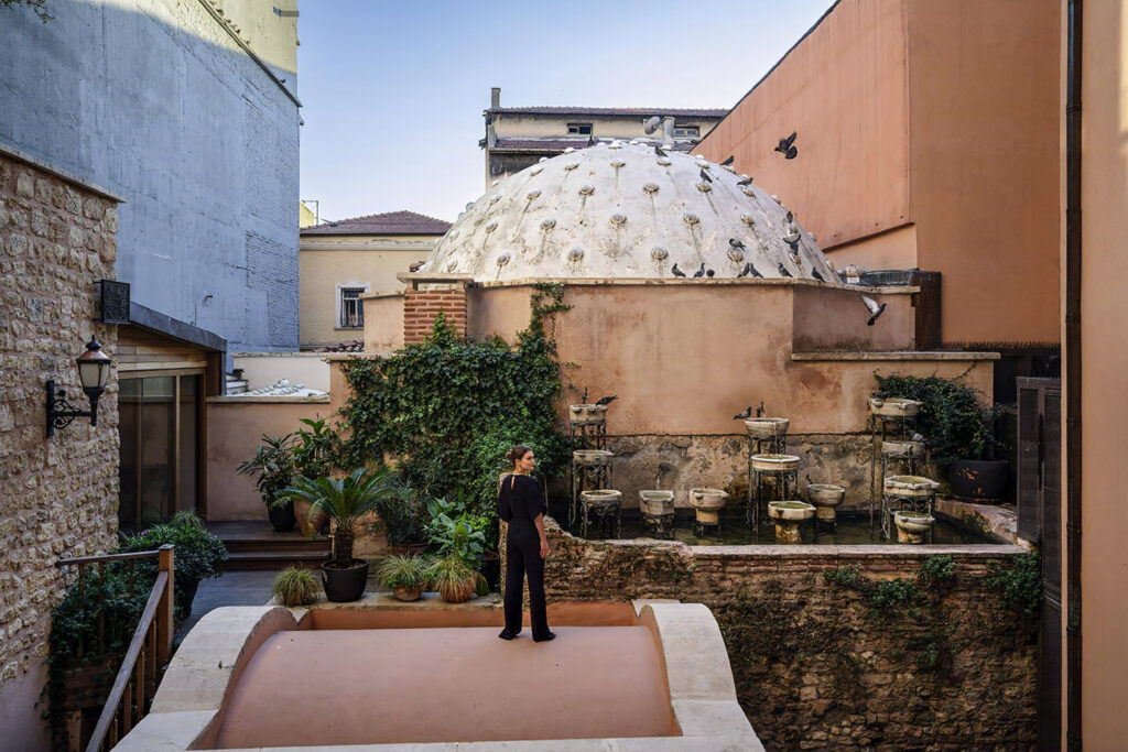 Woman standing beside a preserved Ottoman hammam dome in the MGallery Istanbul hotel courtyard, surrounded by stone basins, ivy, and plants