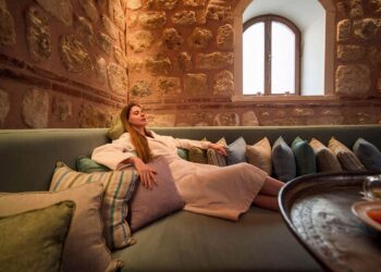 Woman in bathrobe resting on cushioned stone-walled divan in camekan at Tarihi Çeşme Hamamı Karaköy