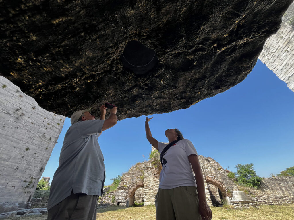 Tour guide photographing the underside of a large contemporary art installation at Yedikule Fortress while a visitor points upward at it, ruined Byzantine brick arches visible behind