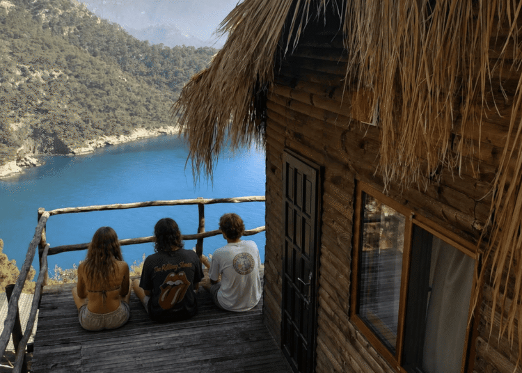 Three travelers relaxing on a wooden terrace overlooking Kabak Bay and the Mediterranean coast near Fethiye, Türkiye.