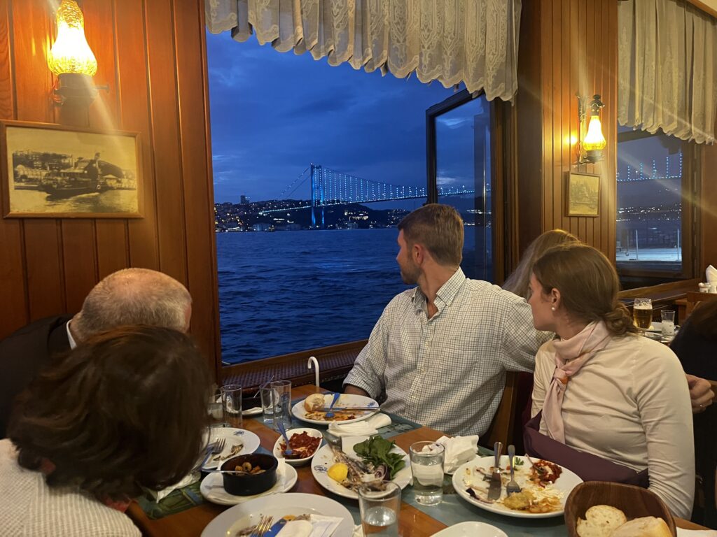 Guests enjoying dinner at a Bosphorus waterfront restaurant with panoramic views of the illuminated Bosphorus Bridge in Istanbul