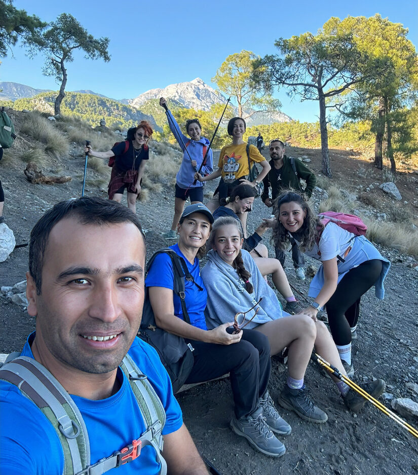 Ali Kütük leading a group of trekkers on a mountain trail with pine trees and a rocky Taurus peak behind, on the Lycian Way near Antalya