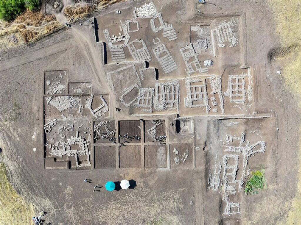 Aerial view of Çayönü Tepesi excavation showing grill-plan stone foundations, with archaeologists and umbrellas below