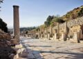 Looking south down Curetes Street, Ephesus, from the upper section — an inscribed column stands in the foreground, statue plinths line the right side, and the Library of Celsus is visible in the far distance