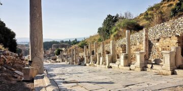 Looking south down Curetes Street, Ephesus, from the upper section — an inscribed column stands in the foreground, statue plinths line the right side, and the Library of Celsus is visible in the far distance