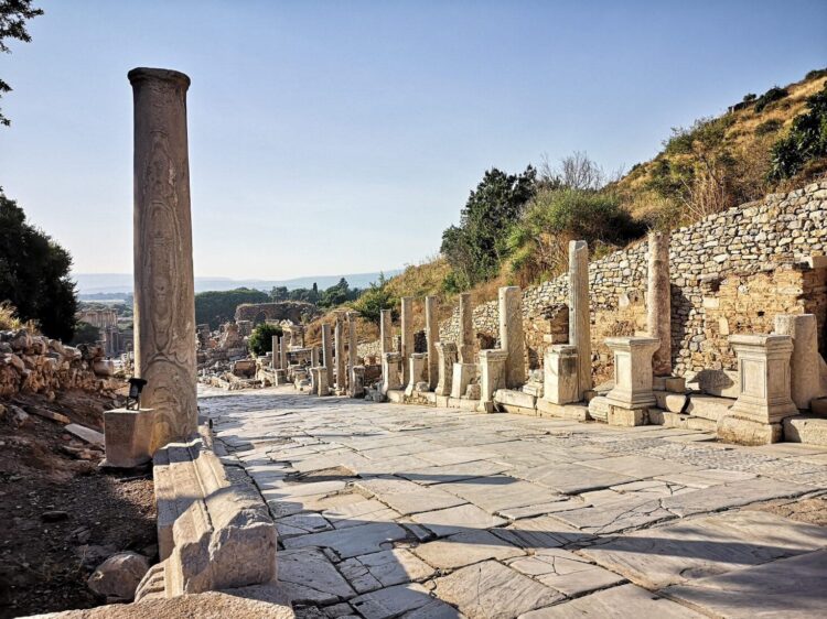 Looking south down Curetes Street, Ephesus, from the upper section — an inscribed column stands in the foreground, statue plinths line the right side, and the Library of Celsus is visible in the far distance