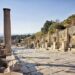 Looking south down Curetes Street, Ephesus, from the upper section — an inscribed column stands in the foreground, statue plinths line the right side, and the Library of Celsus is visible in the far distance