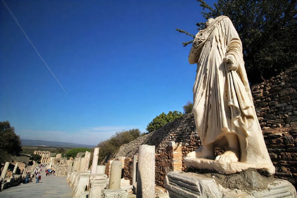 A headless marble statue in a Roman toga stands on its plinth along Curetes Street, Ephesus, with the full sweep of the marble-paved street and the Library of Celsus visible far below