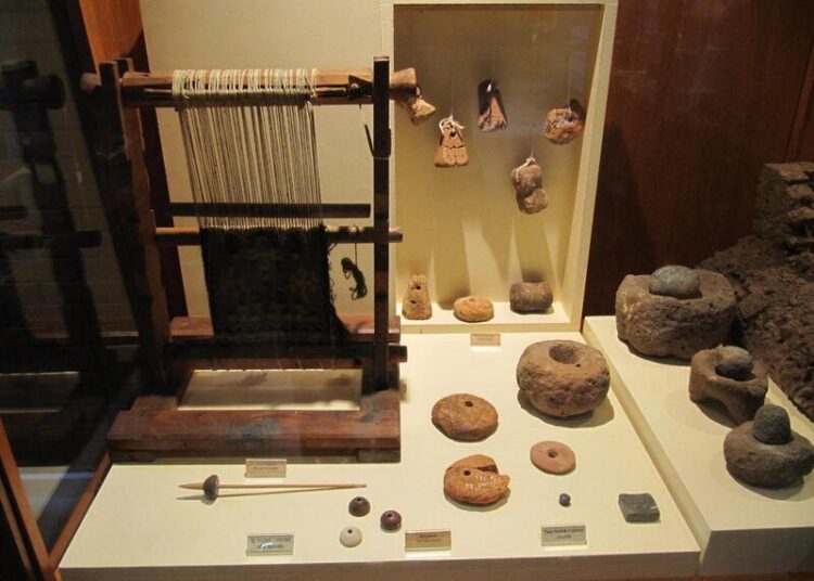 Museum display featuring a reconstructed ancient loom alongside spindle whorls, stone weaving tools, and textile production artifacts.