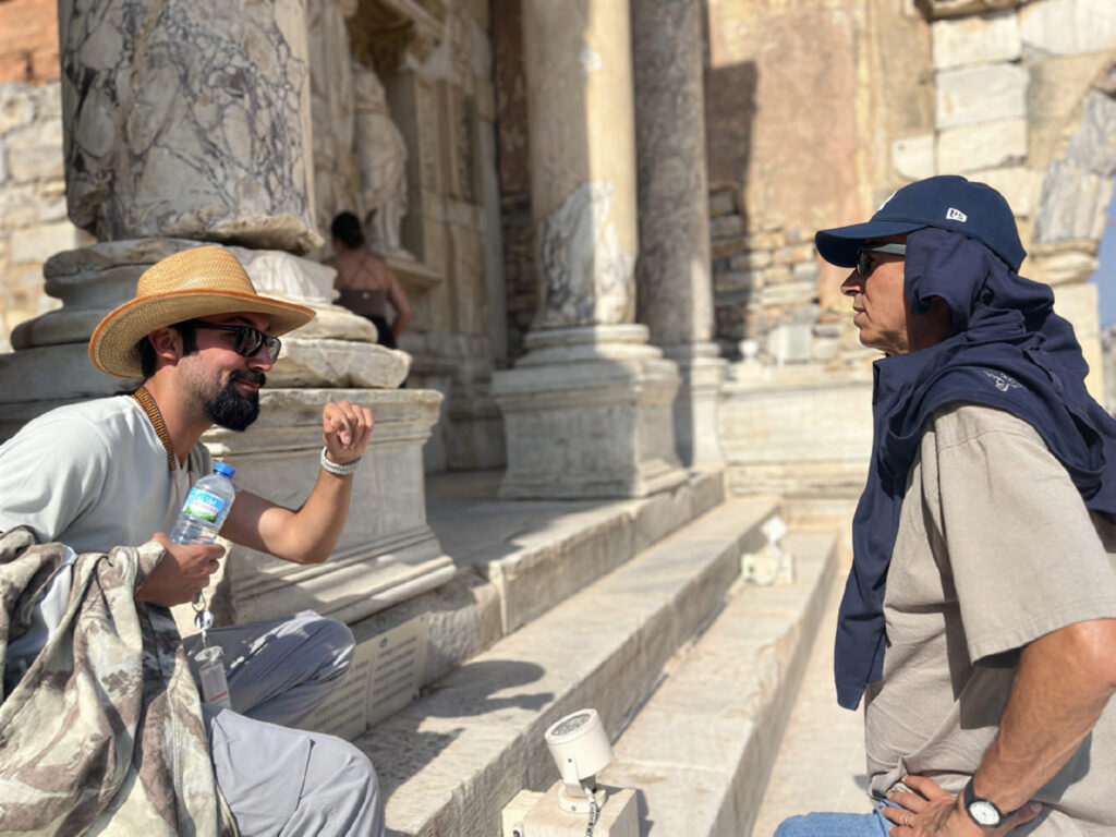 A licensed Ephesus guide in a straw hat talks through a monument with a guest on the marble steps of the Temple of Hadrian on Curetes Street — the kind of stop an unguided eleven-minute walk skips entirely