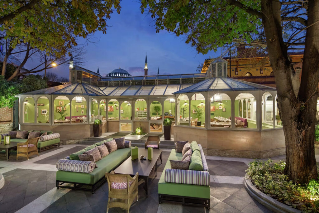 Sofia Terrace glass conservatory restaurant at Hagia Sofia Mansions at dusk, with Sultanahmet mosque dome and minarets rising above the roofline, outdoor lounge seating in foreground