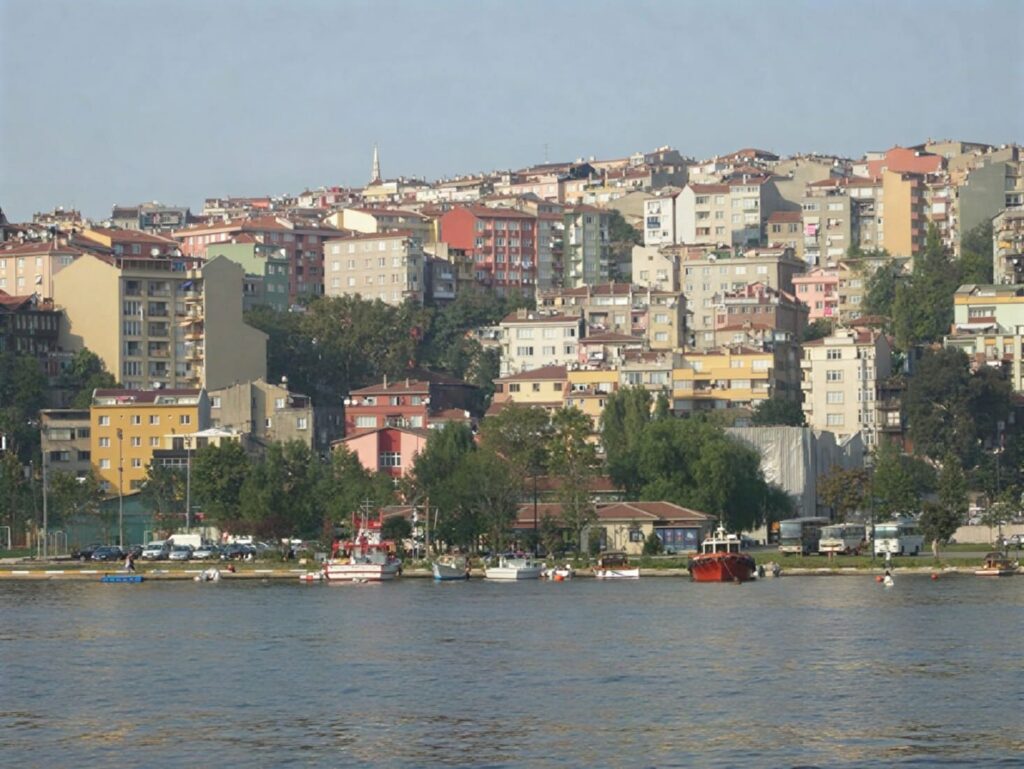 Hasköy seen from the Golden Horn: densely packed apartment buildings in warm afternoon light climb a steep hillside above the waterfront, with small boats moored along the shore below