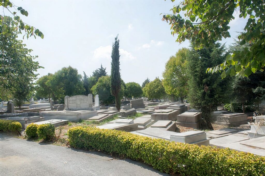 A paved path through a manicured section of the Hasköy Jewish Cemetery in Istanbul, lined with cypress trees, trimmed hedges, and stone grave markers under a summer sky