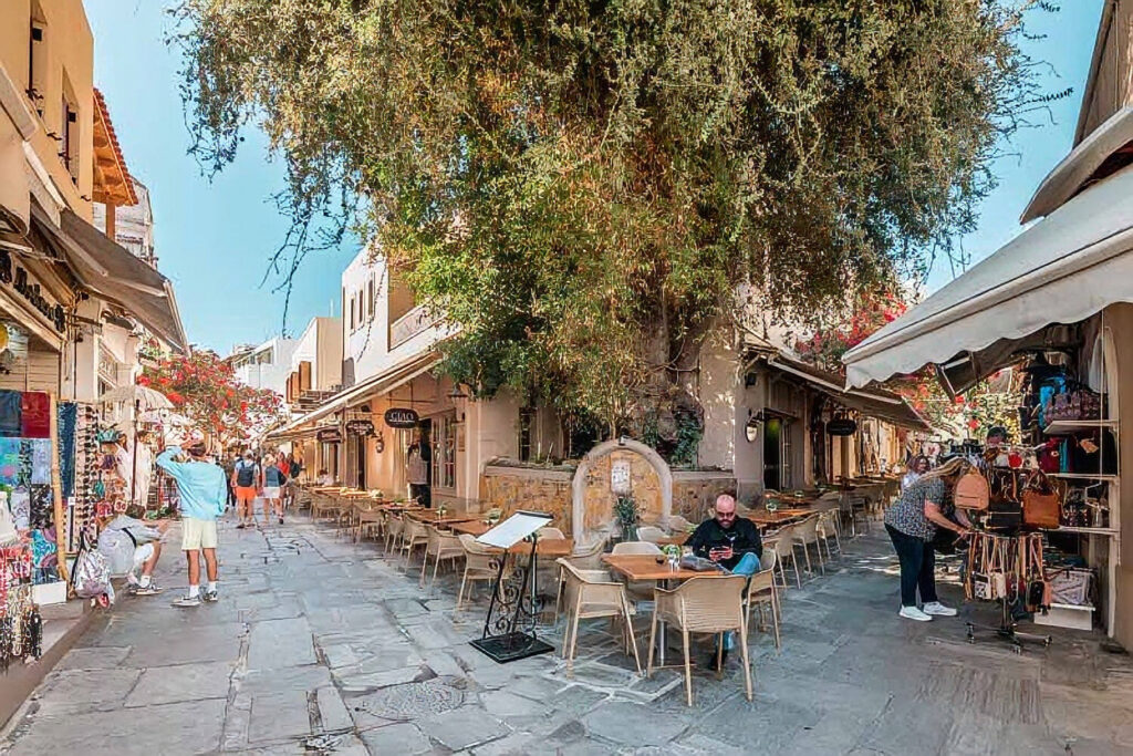 A shaded café street in Kos Old Town beneath a wide plane tree — five minutes on foot from the ferry terminal where boats arrive from Bodrum and Turgutreis