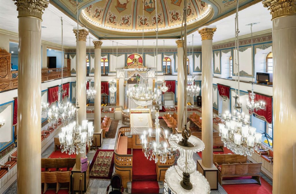 Interior of the Maalem Synagogue in Hasköy, Istanbul, viewed from the upper gallery: tall white Corinthian columns, a painted dome with medallion frescoes, multiple crystal chandeliers, a central ornate bimah, and red Star of David curtains — the synagogue dates to 1754