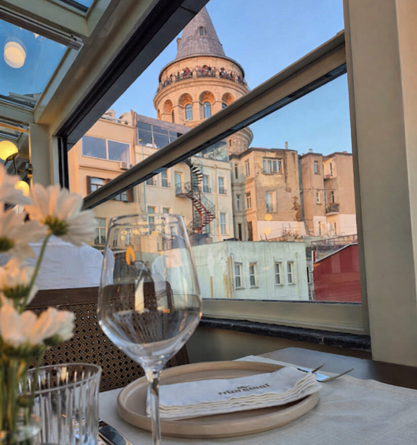 Table set with Manifest-branded linen and wine glass with Galata Tower rising through the open glass panel at golden hour