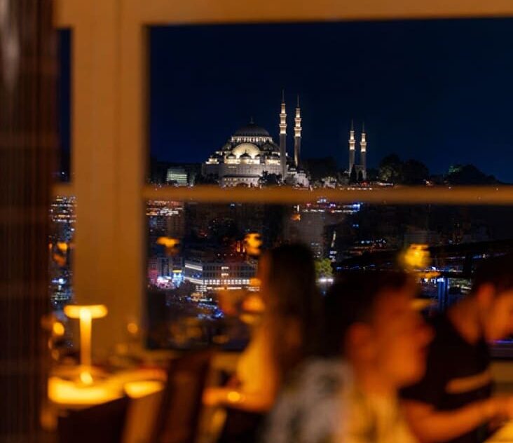 Diners at Manifest Roof Restaurant at night with the illuminated Süleymaniye Mosque and Golden Horn visible through the panoramic windows