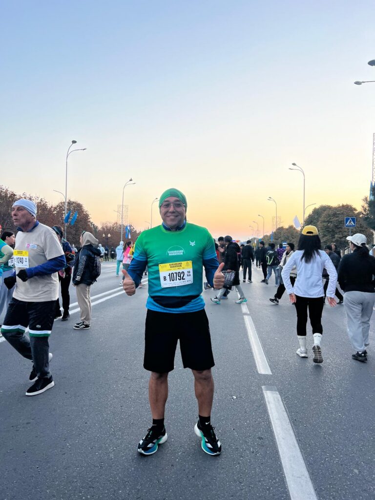 Orçun Kamit standing on a closed city street before the start of the Samarkand Marathon, wearing a blue running shirt and race bib while giving two thumbs up.