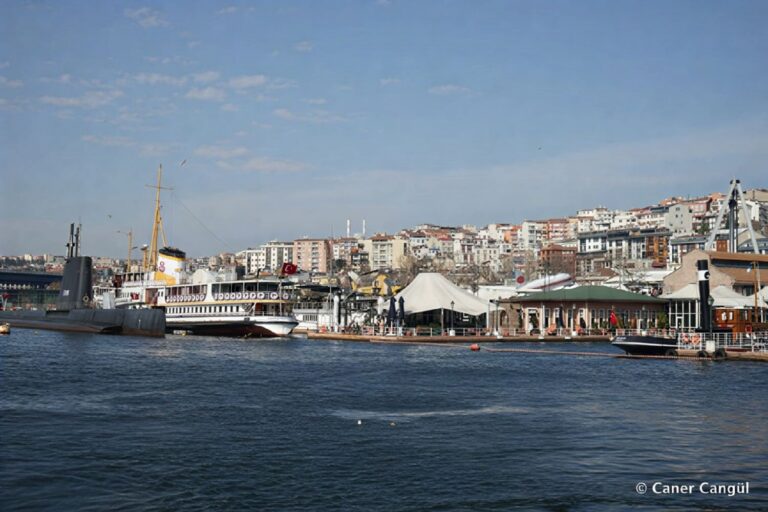 The Rahmi M. Koç Museum waterfront in Hasköy, Istanbul, seen from the Golden Horn: the TCG Uluçalireis submarine and Fenerbahçe ferry moored side by side, with white museum canopies, the shore café, and the residential hillside rising behind