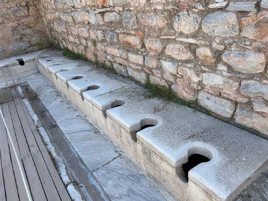 The 36-seat Roman public latrines beside the Scholastikia Baths on Curetes Street, Ephesus — a row of marble toilet seats with keyhole openings set over a continuously flushed water channel
