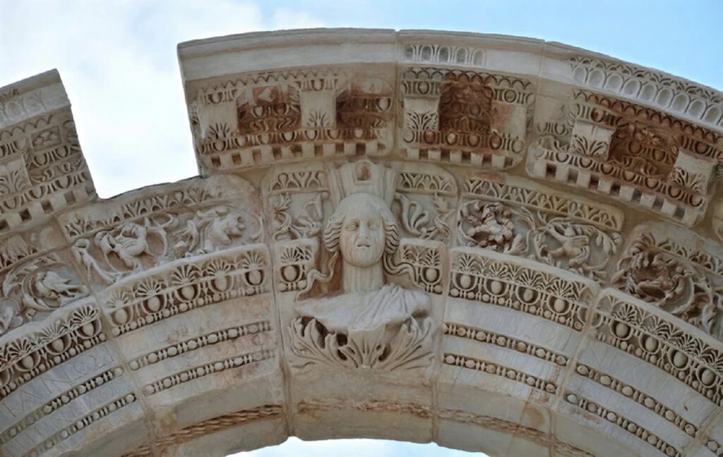 Close-up of the Medusa (or Tyche) head at the keystone of the Temple of Hadrian's inner arch on Curetes Street, Ephesus, surrounded by intricate acanthus scrolls and mythological figures — the detail most visitors walk past without looking up