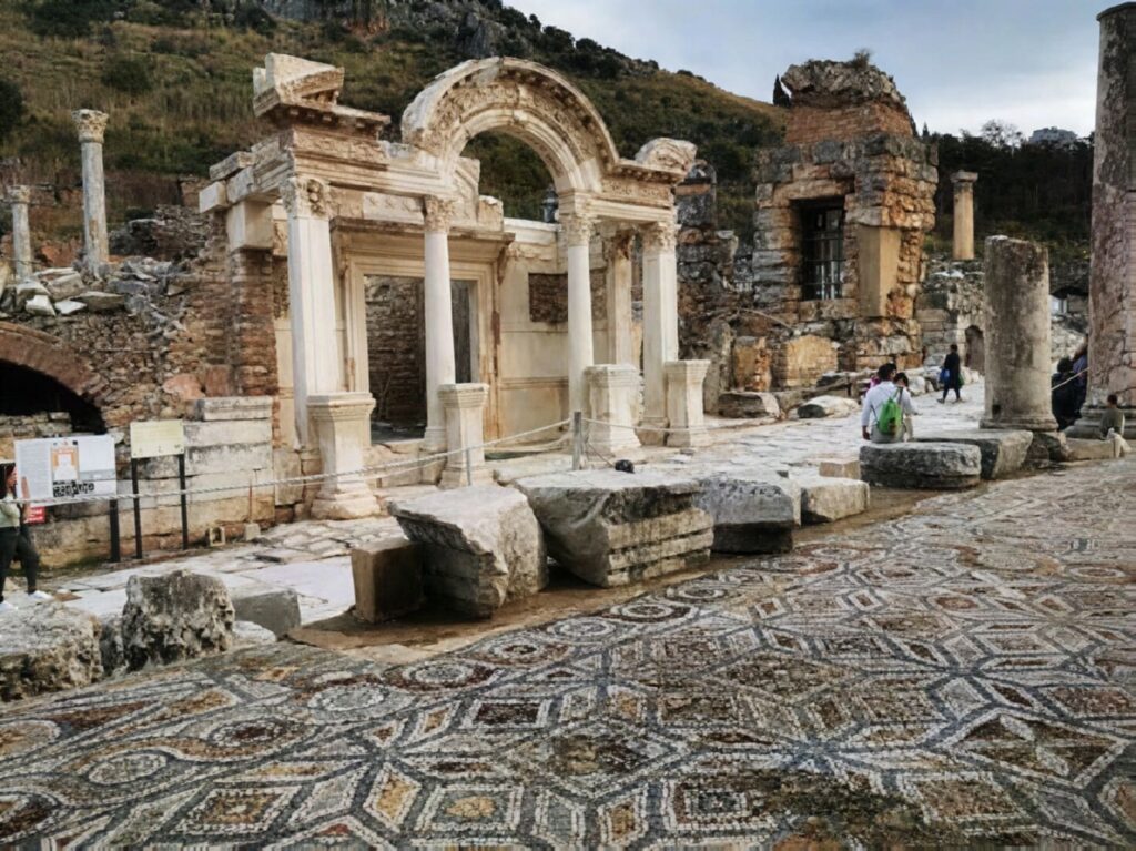 The Temple of Hadrian viewed from street level on Curetes Street, Ephesus, with an elaborate geometric Roman mosaic filling the foreground — one of the most photographed spots in the ancient city