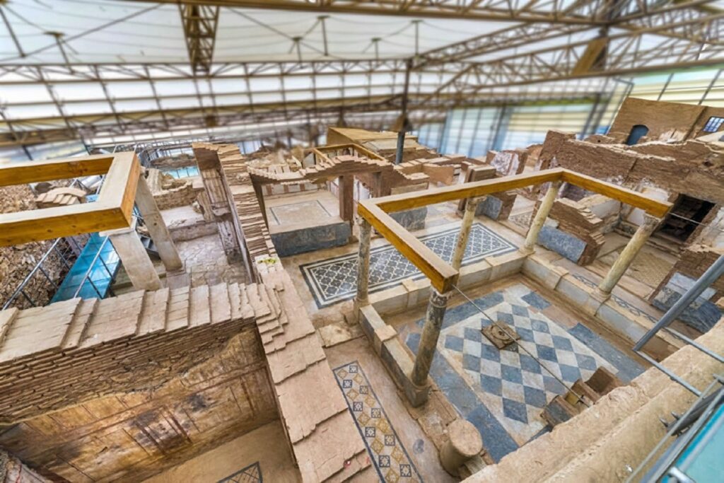 Aerial view of the Terrace Houses of Ephesus under their modern steel and glass conservation shelter, revealing multiple interconnected rooms with geometric mosaic floors, multi-storey brick walls and peristyle courtyards