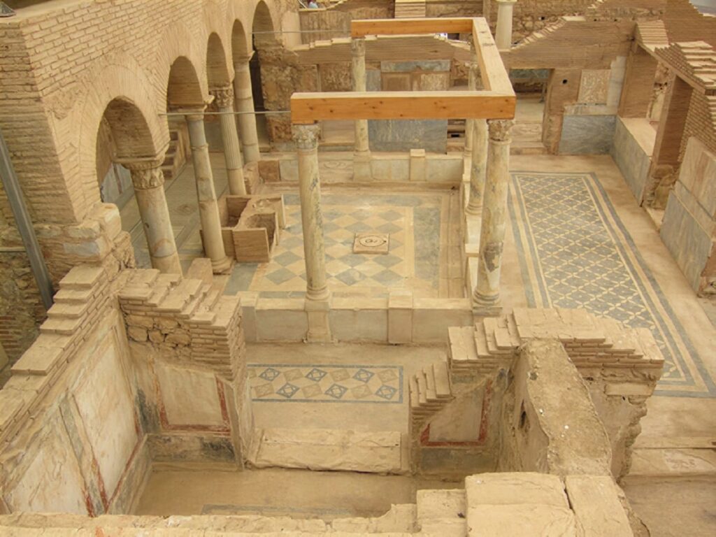 Bird's-eye view into a Terrace House courtyard in Ephesus, showing a preserved diamond-pattern mosaic floor, Corinthian columns and Roman brick arches photographed during an early phase of excavation