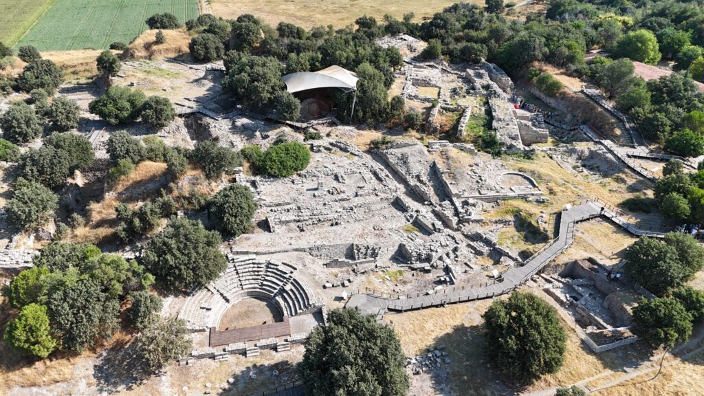 Aerial view of the archaeological site of Troy at Hisarlık, showing the excavated ruins, ancient theater, stone walls, and walking paths.