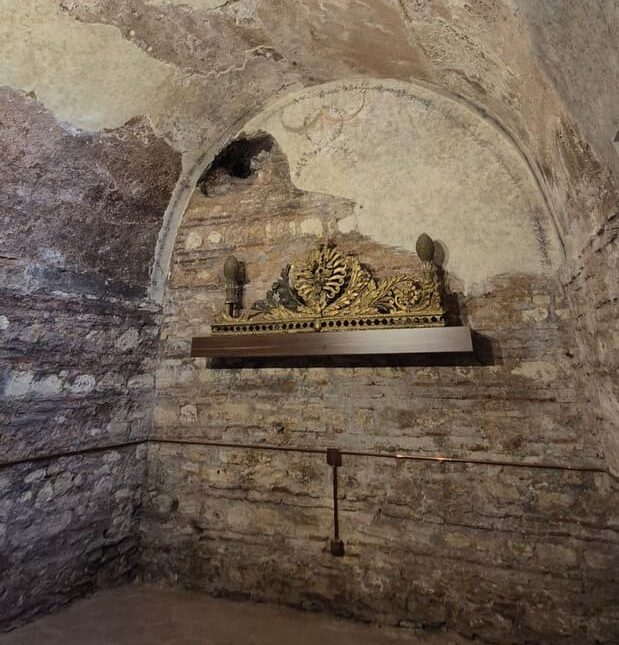 A rough stone vaulted chamber beneath Yanbol Synagogue with faded frescoes, a skylight, and an ornate gilded Torah ark crown displayed in a niche