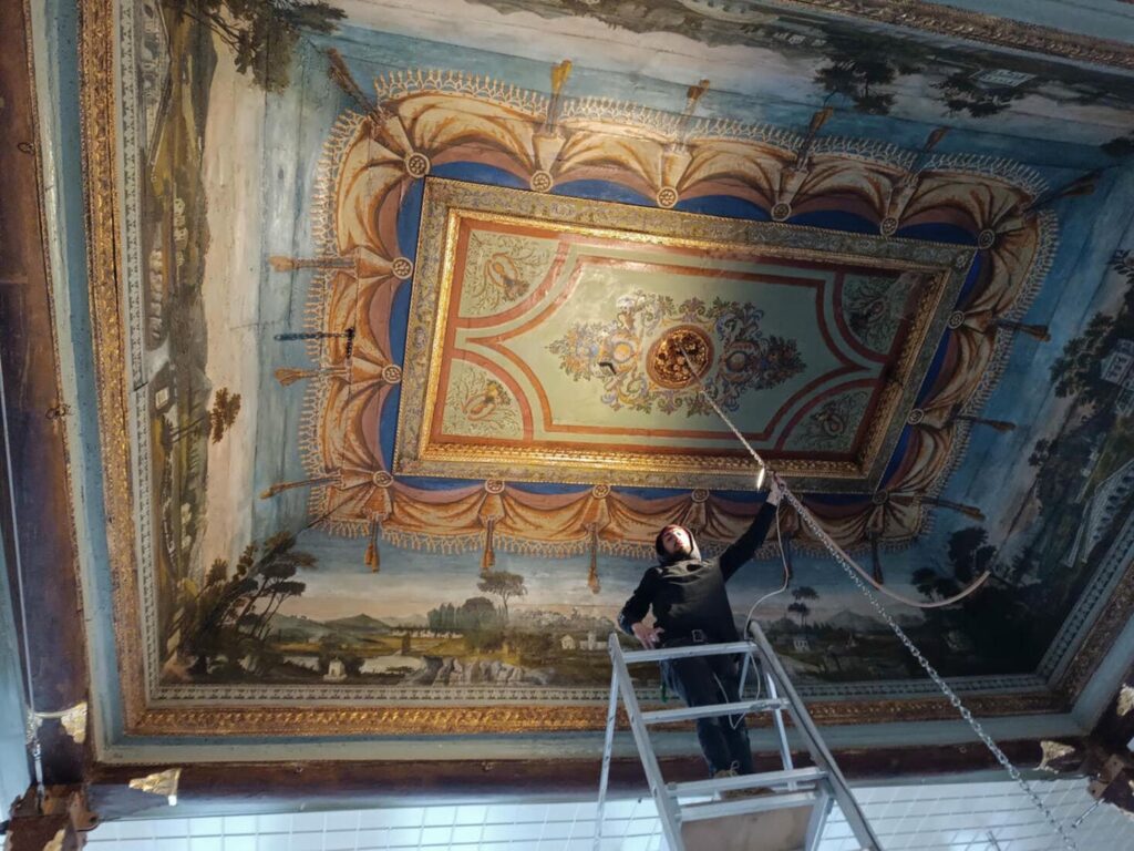 A conservator on a ladder carefully restoring the painted ceiling of Yanbol Synagogue, showing landscape murals and ornate baroque framing