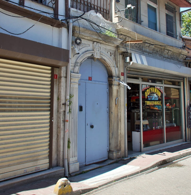 The unassuming street facade of Yanbol Synagogue in Balat, Istanbul, featuring an ornate carved stone arched doorway between a shuttered shop and a tobacco store