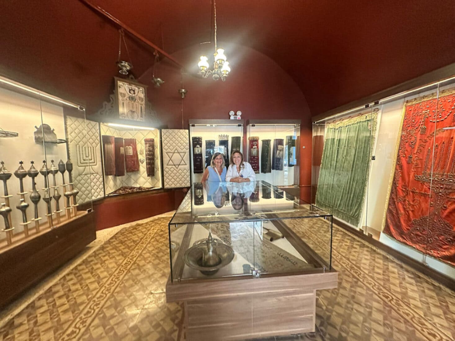 Two women standing inside the Yanbol Synagogue's Judaica museum room, surrounded by Torah mantles, silver ritual objects, and embroidered textiles in display cases