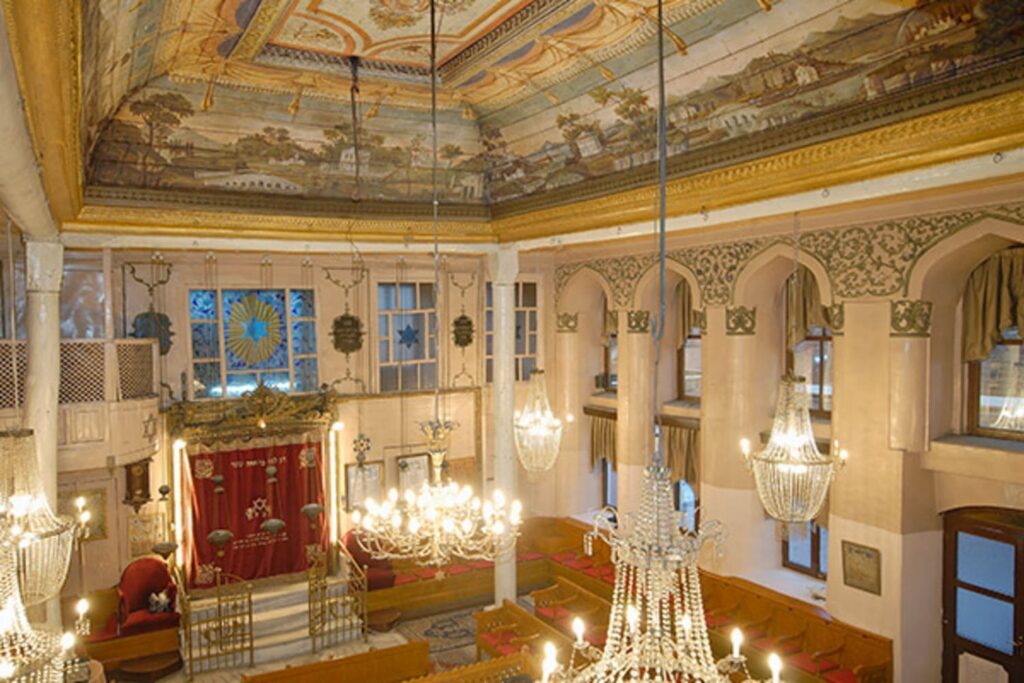 Wide panoramic view of Yanbol Synagogue's main sanctuary showing the full painted ceiling with landscape murals, gilded cornices, crystal chandeliers, white arched walls, and the Torah ark at the far end