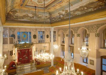 Wide panoramic view of Yanbol Synagogue's main sanctuary showing the full painted ceiling with landscape murals, gilded cornices, crystal chandeliers, white arched walls, and the Torah ark at the far end