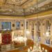 Wide panoramic view of Yanbol Synagogue's main sanctuary showing the full painted ceiling with landscape murals, gilded cornices, crystal chandeliers, white arched walls, and the Torah ark at the far end