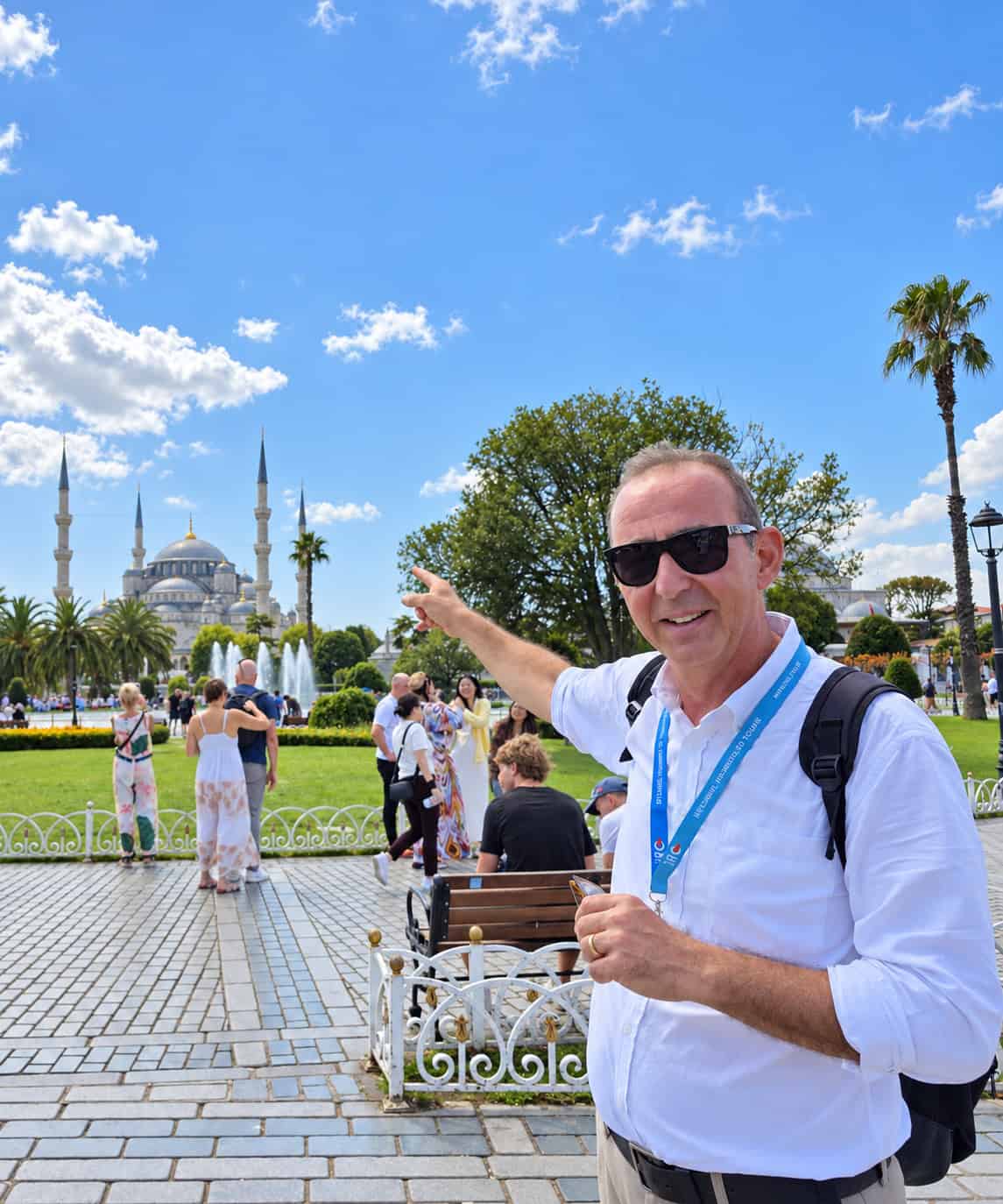 Ahmet Can Gürel, his guide card on a lanyard, pointing across Sultanahmet Square toward the Blue Mosque