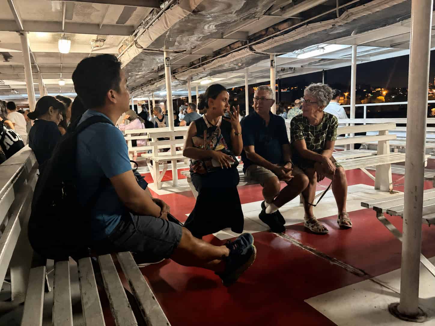 A group of guests sits with their guide on the open deck of a public ferry at dusk